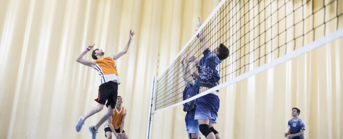 Das Foto zeigt zwei Teams beim Volleyball. Ein Mann springt hoch, um den Ball zu spielen.