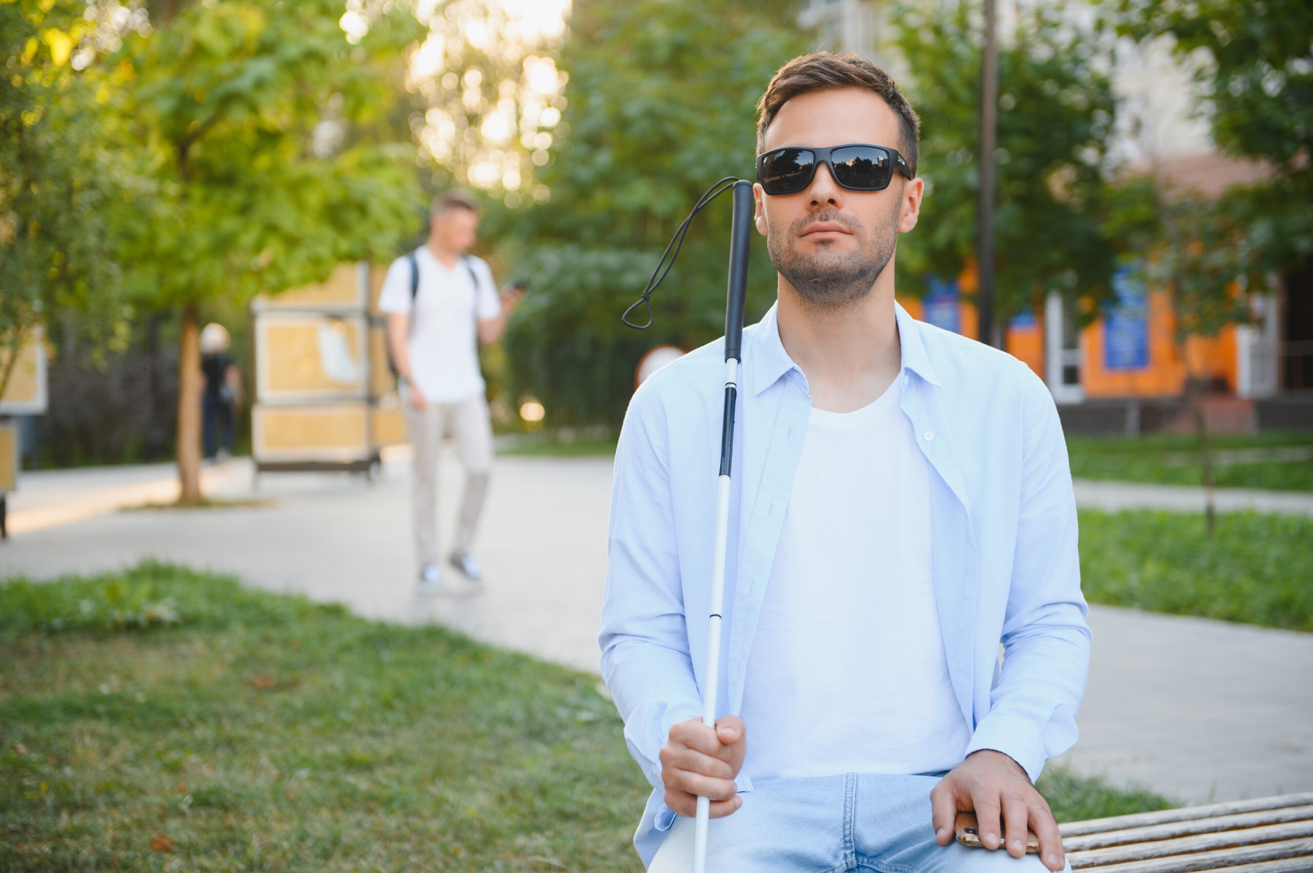 Ein blinder Mann mit Sonnenbrille und Blindenstock sitzt auf einer Parkbank.