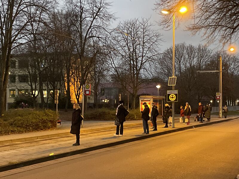 Bild von einer Straße, Menschen stehen an der Haltestelle